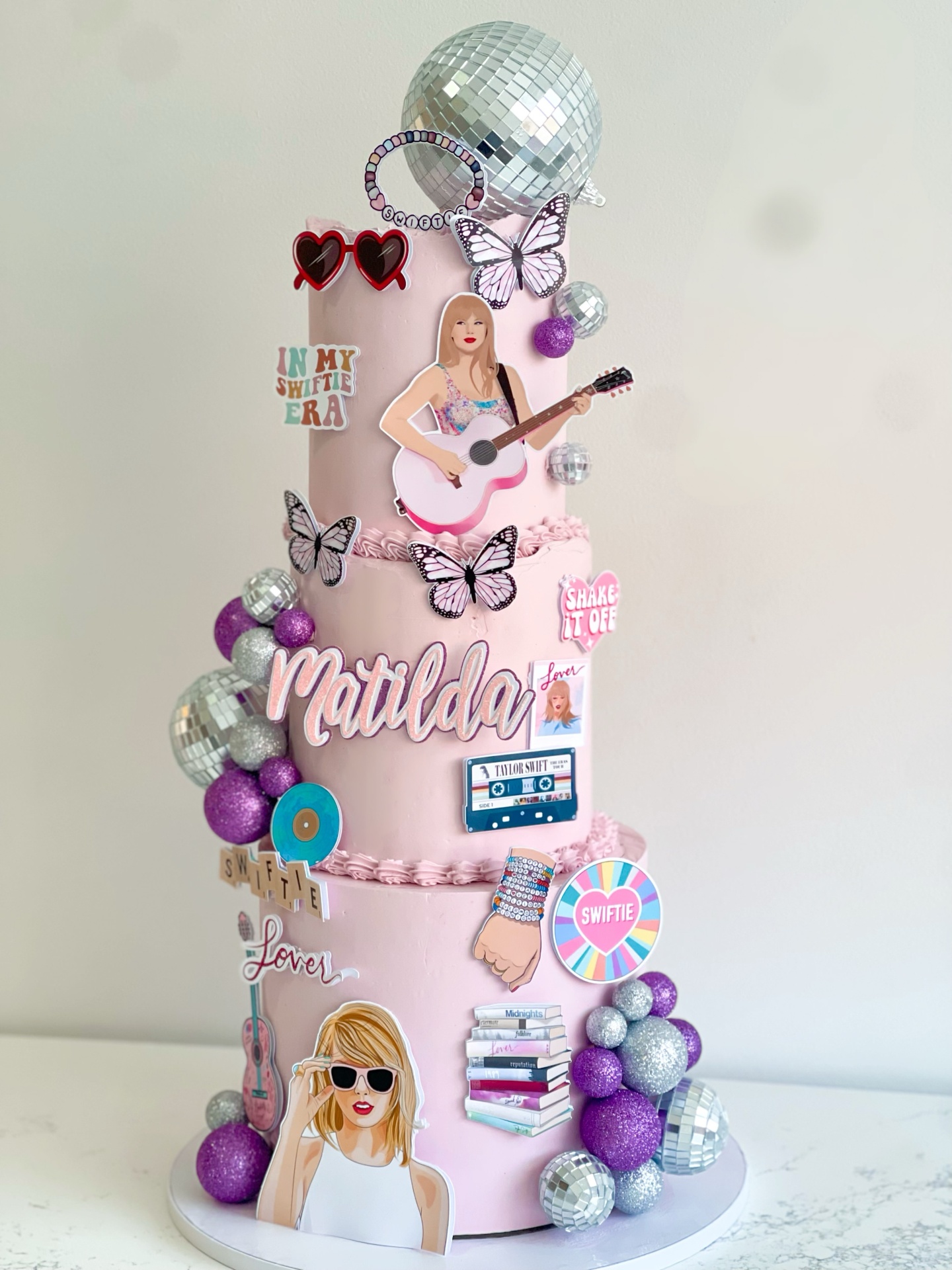 Three-tier pink Taylor Swift Swiftie themed birthday cake with disco ball, butterflies and album decorations